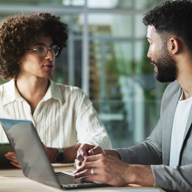 Twee mannen zitten aan een bureau en kijken naar elkaar, laptop staat op tafel.