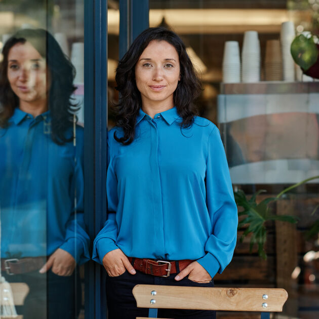 Vrouw met handen in de zakken en blauwe blouse aan leunt tegen de deur.