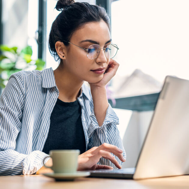 Vrouw met bril op zit aan tafel en kijkt denkend naar laptop.