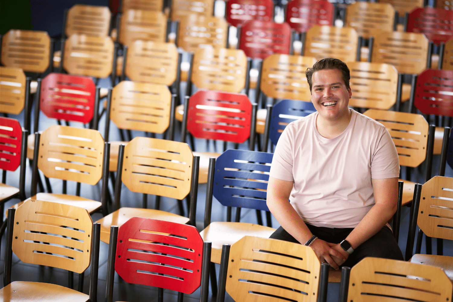 Man in roze shirt zit op schoolstoel met schoolstoelen om zich heen
