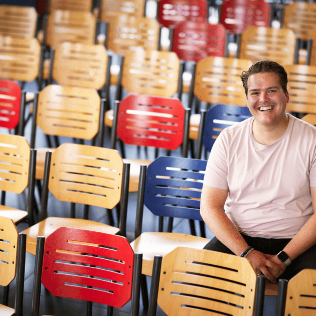 Man in roze shirt zit op schoolstoel met schoolstoelen om zich heen