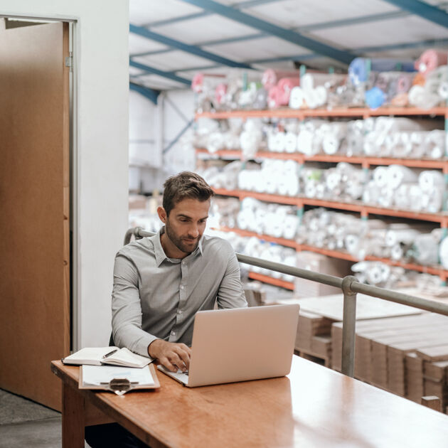 Man met een blouse aan zit achter een laptop in een hal. 