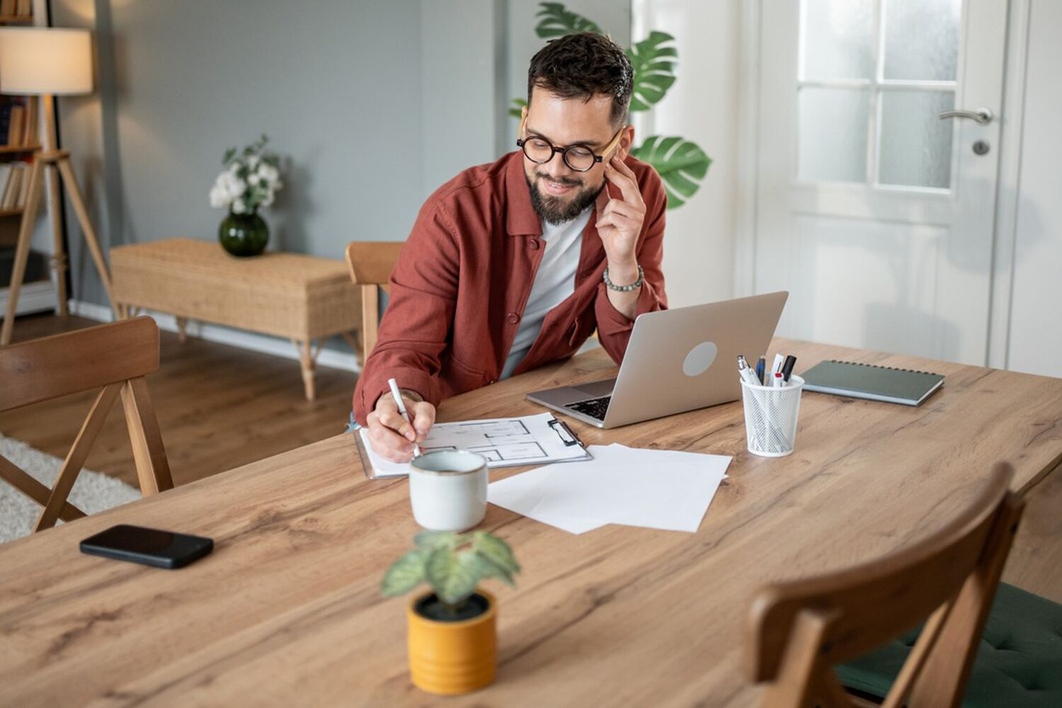 man-met-bril-achter-laptop-schrijft-op-kladblok-header Zittende man met bril en rode blouse schrijft in een notitieblok naast zijn laptop.