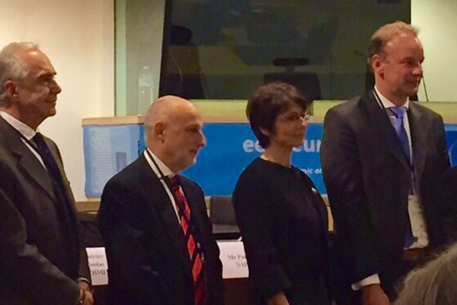 Een rij van 8 net geklede mensen staan in een persconferentie zaal 