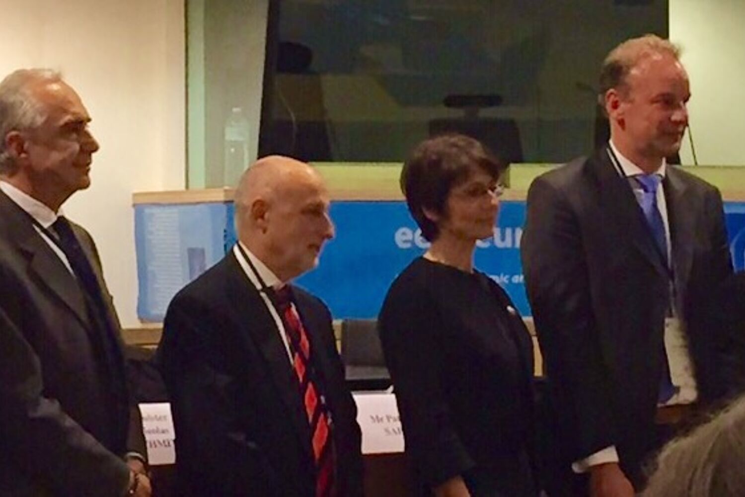 Een rij van 8 net geklede mensen staan in een persconferentie zaal 