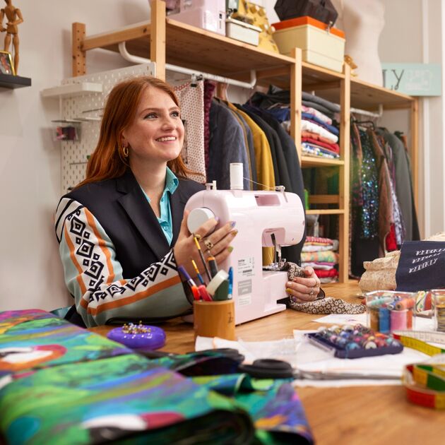 Lachende vrouw zit aan tafel met een naaimachine en kijkt dromend weg. 