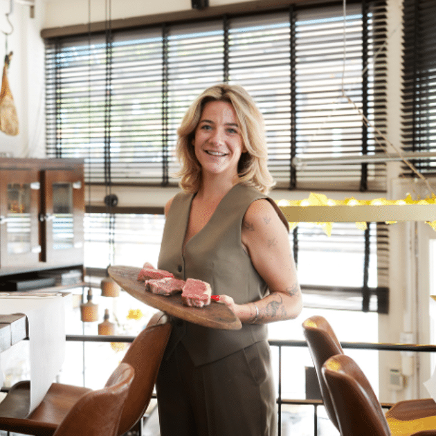 Vrouw met groen jasje houdt houten plank vast met vlees in restaurant 