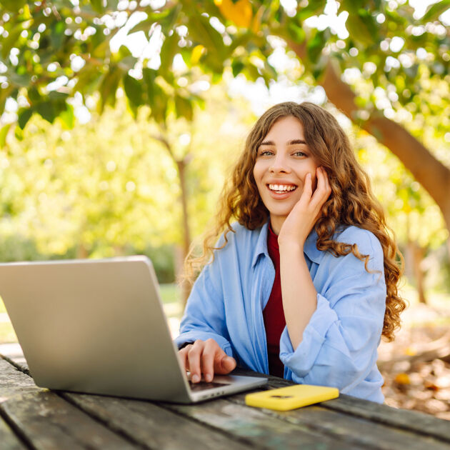 Vrouw zit buiten achter laptop en kijkt lachend in de camera.