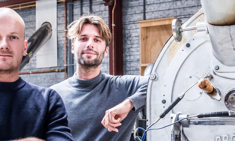 Twee mannen in donkere shirts één heeft zijn armen gevouwen en de andere leunt tegen machine