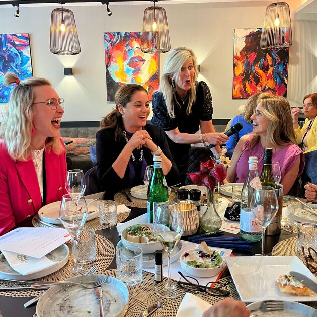 Vrouw staat met microfoon bij andere vrouwen aan tafel. 