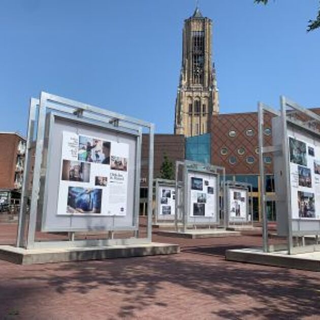 Buiten op een plein staan grote borden op betonnen blokken. Op deze grijze borden staan foto's en verhalen. Er is een kerkklok op de achtergrond.