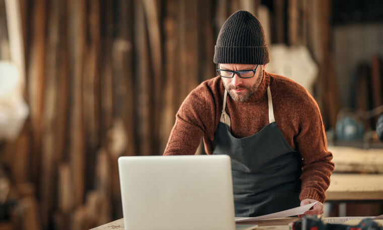Man in schort en beanie kijkt naar laptop in houtbewerking studio