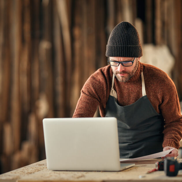 Man in schort en beanie kijkt naar laptop in houtbewerking studio