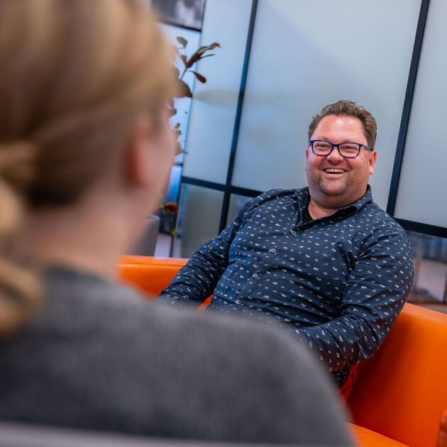 Man in blauw shirt en bril zit op een oranje bank en praat met iemand.