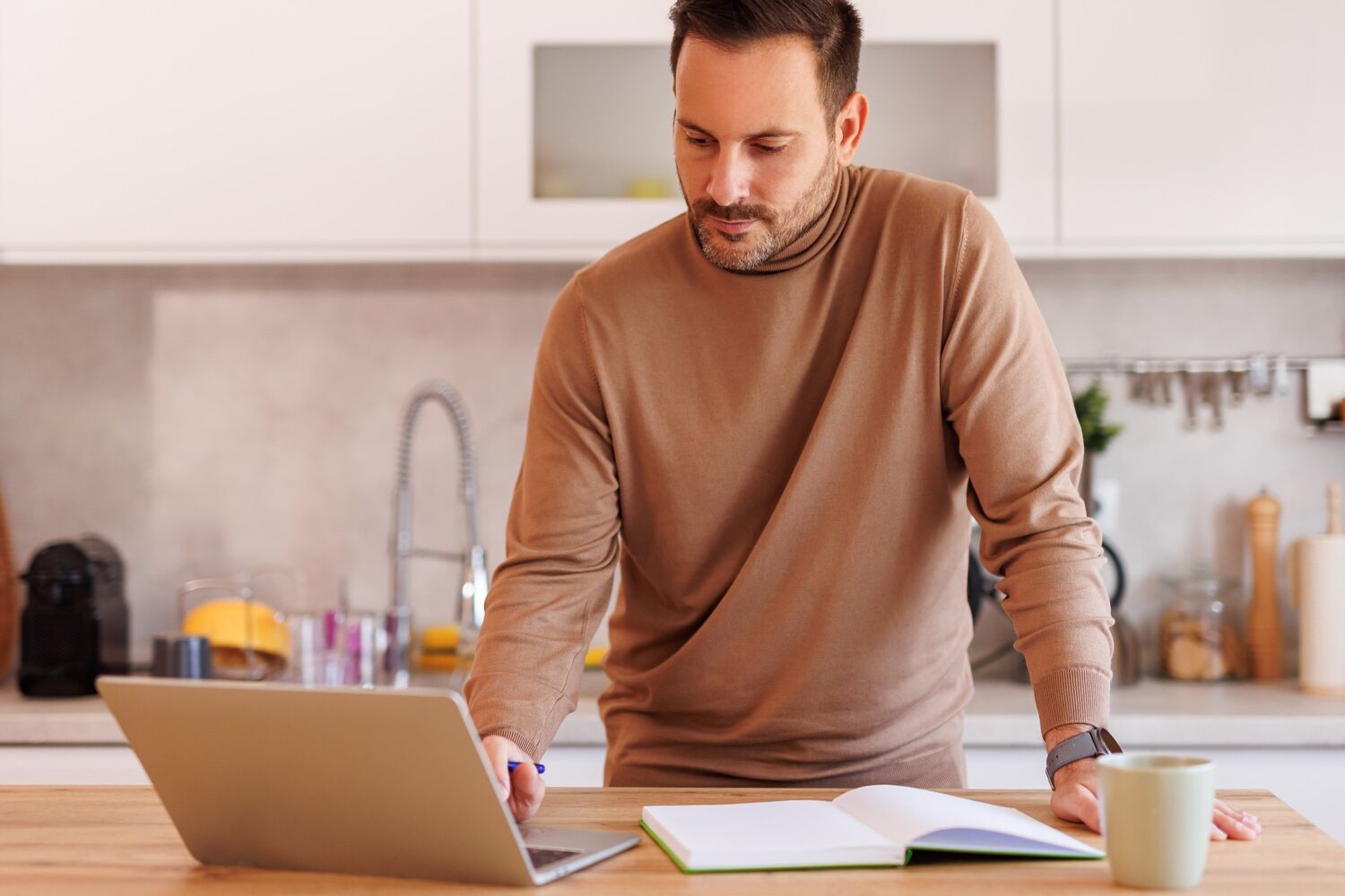 Man staat achter het aanrecht en kijkt serieus naar laptop.