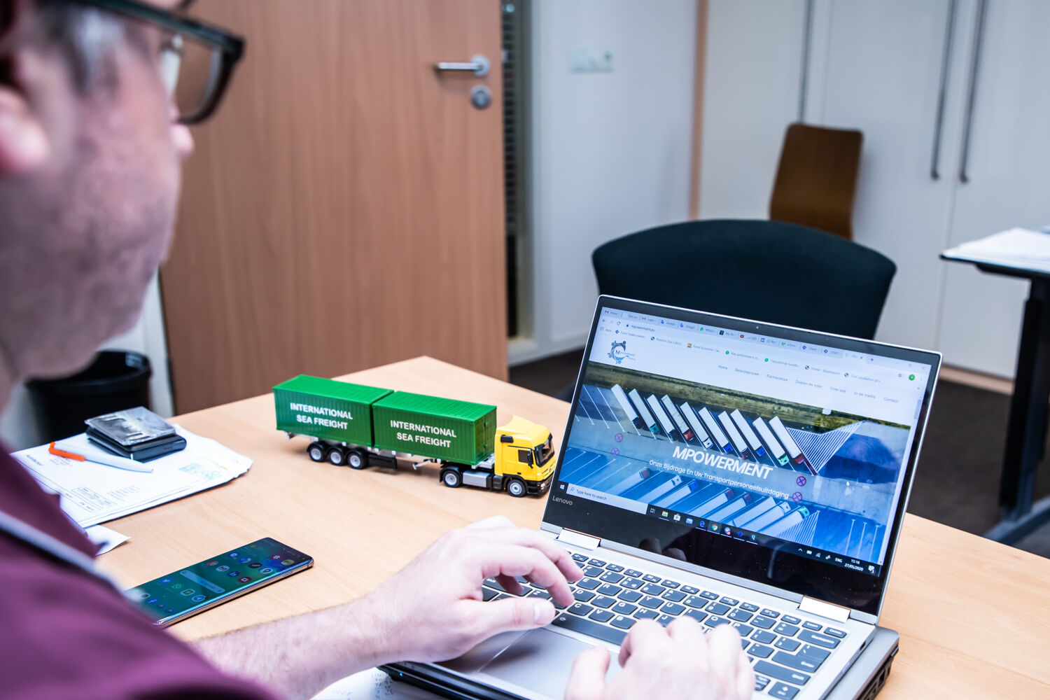 Man typt op laptop met scherm naar camera en een groene speelgoed truck op tafel