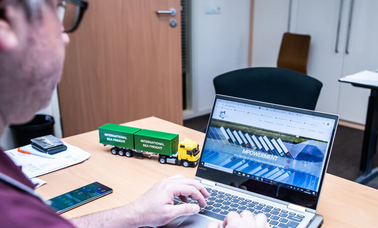 Man typt op laptop met scherm naar camera en een groene speelgoed truck op tafel
