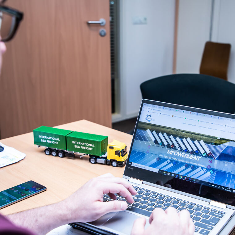 Man typt op laptop met scherm naar camera en een groene speelgoed truck op tafel