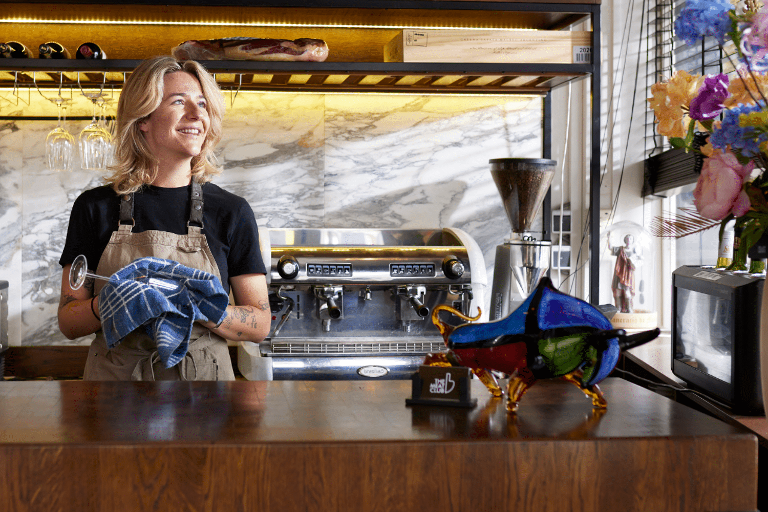 Een blonde vrouw staat achter een houten bar en poetst een wijnglas terwijl ze lacht.