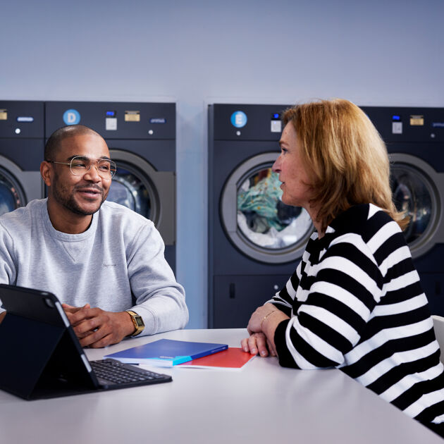 Een man en een vrouw zijn zittend aan het praten, voor wasmachines met een tablet op tafel.