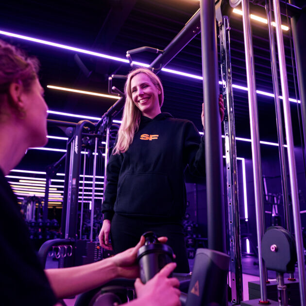 Een vrouw zit, andere vrouw staat. Ze kijken lachend naar elkaar in sportschool.