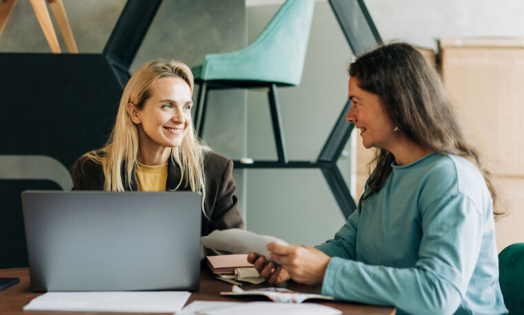 Twee vrouwen zitten aan tafel, met de laptop op tafel. Ze kijken elkaar lachend aan. 