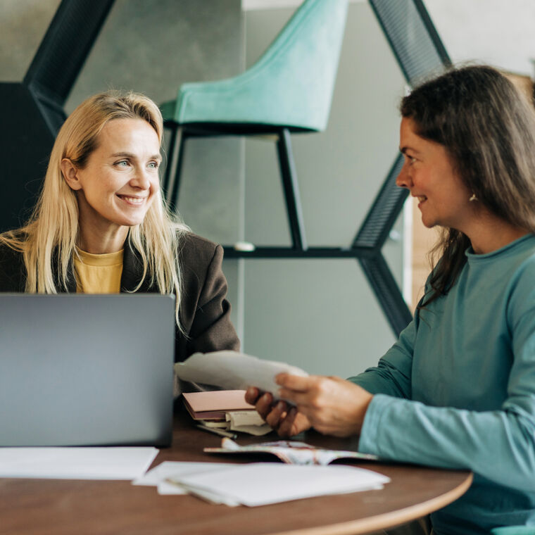 Twee vrouwen zitten aan tafel, met de laptop op tafel. Ze kijken elkaar lachend aan. 