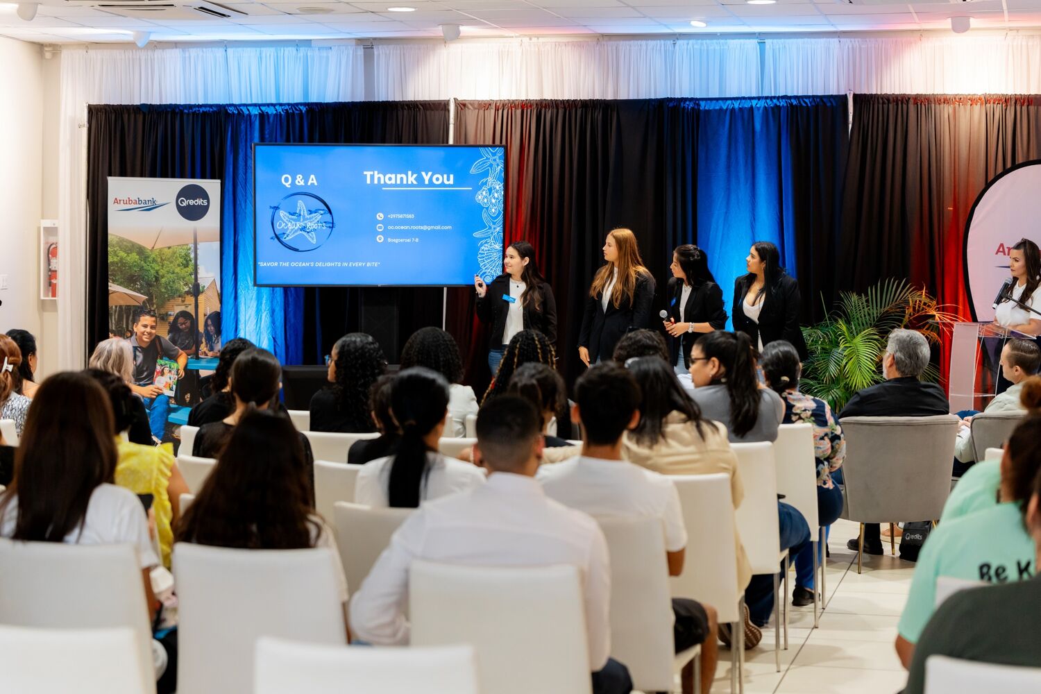 Be-Your-Own-Boss-Pitch-Event-Pitches Een zaal met witte stoelen waar mensen op zitten terwijl ze kijken naar een groep vrouwen die iets aan het pitchen zijn bij een elektrisch scherm