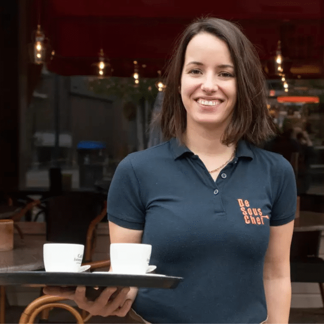 Vrouw met dienblad in handen staat lachend voor restaurant.