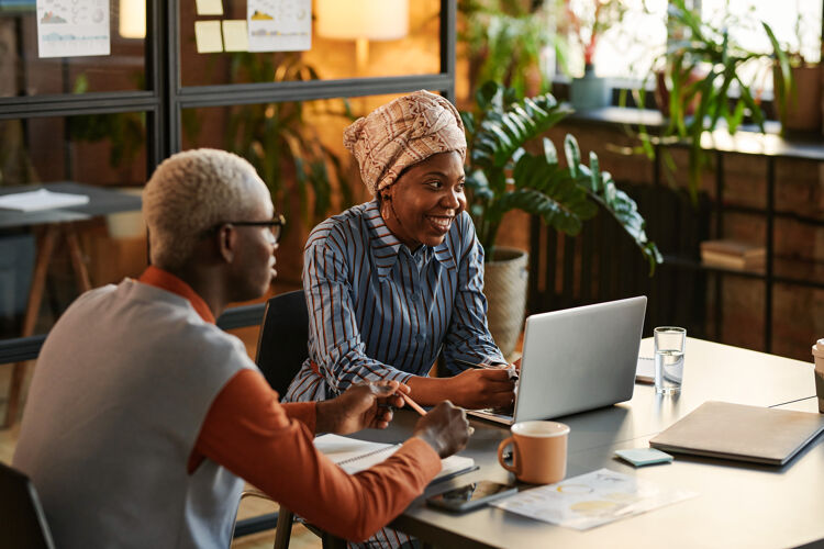 Vrouw met hoofddoek en blauwe blouse lacht zit achter laptop man ernaast