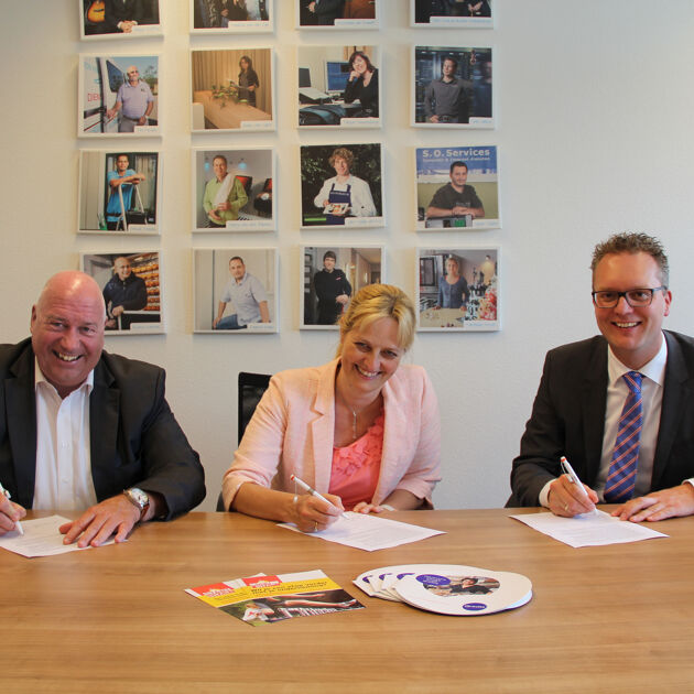 Drie lachende mensen zitten aan een houten tafel en ondertekenen een papier. Er hangen foto's aan de muur