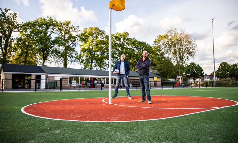 Op de foto staan twee mannen in de midden cirkel van een korfbalveld. Ze staan naast een korfbal paal en kijken naar de camera. Een van de mannen heeft een korfbal vast. 