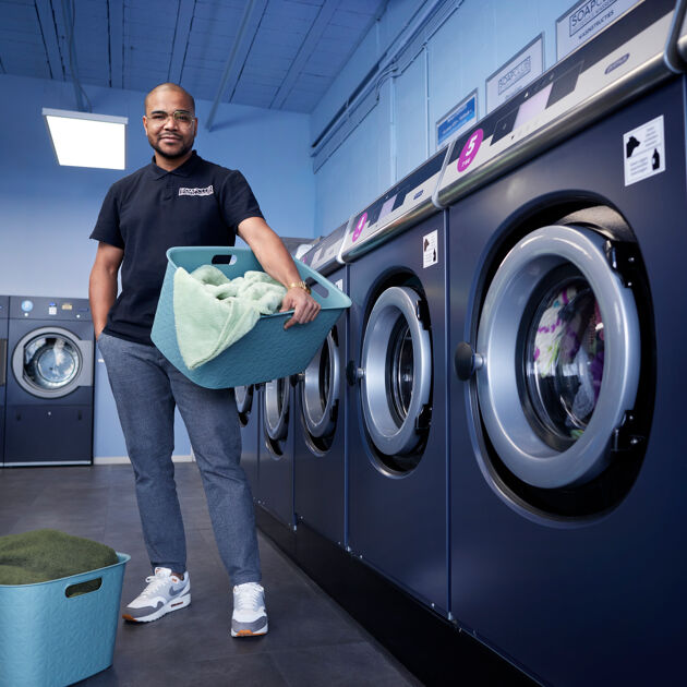 Een man staat met een wasmand met was erin naast wasmachines.