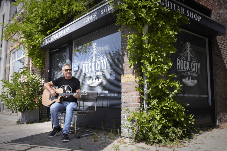 Man in zwart shirt en zonnebril zit buiten gitaar te spelen bij een raam met 'rock city' erop geschreven