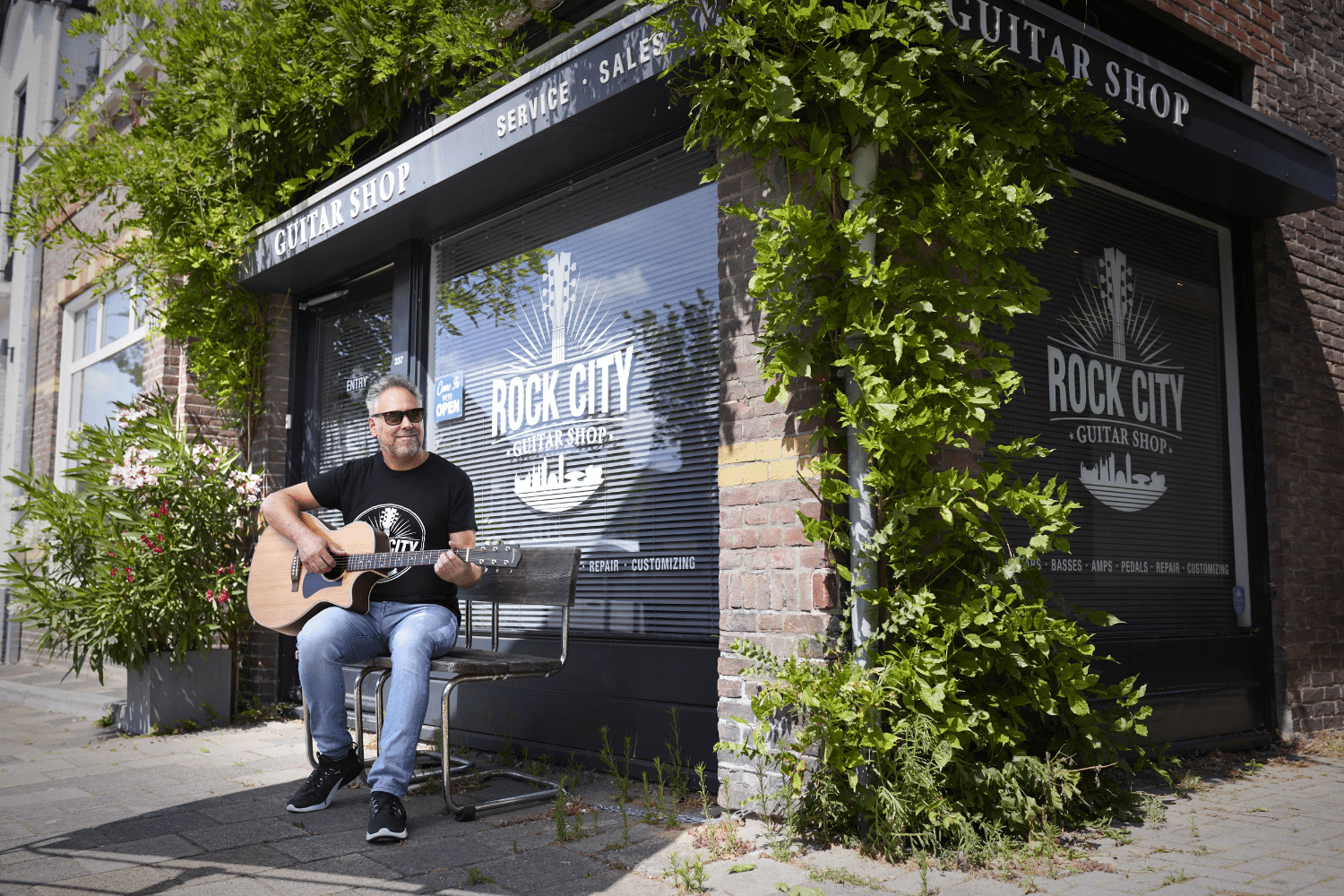 Man in zwart shirt en zonnebril zit buiten gitaar te spelen bij een raam met 'rock city' erop geschreven