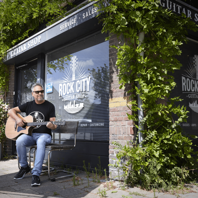 Man in zwart shirt en zonnebril zit buiten gitaar te spelen bij een raam met 'rock city' erop geschreven