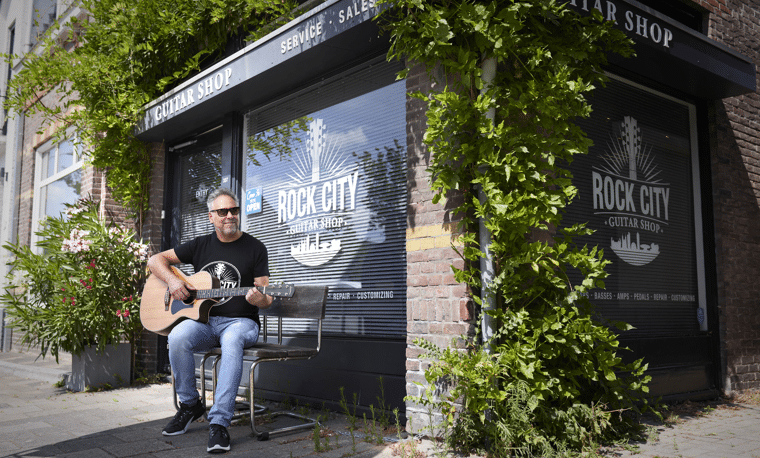 Man in zwart shirt en zonnebril zit buiten gitaar te spelen bij een raam met 'rock city' erop geschreven