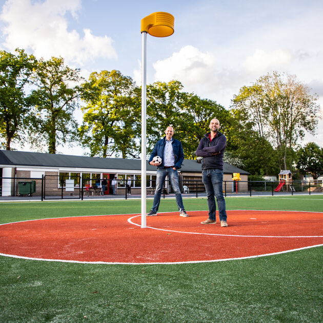 Op de foto staan twee mannen in de midden cirkel van een korfbalveld. Ze staan naast een korfbal paal en kijken naar de camera. Een van de mannen heeft een korfbal vast. 