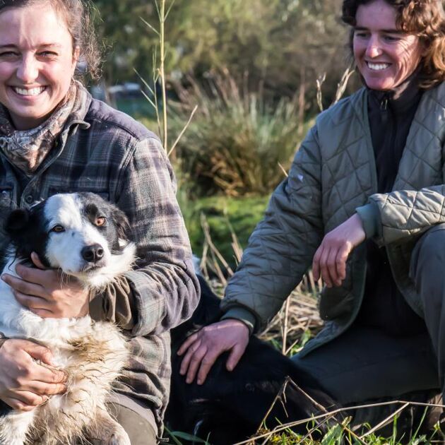 Twee vrouwen buiten met een hond zitten op het gras en lachen.