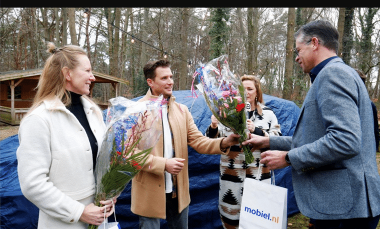 Vier mensen staan buiten bij veel bomen en bij een blauwe auto. Twee mensen hebben bloemen vast. 