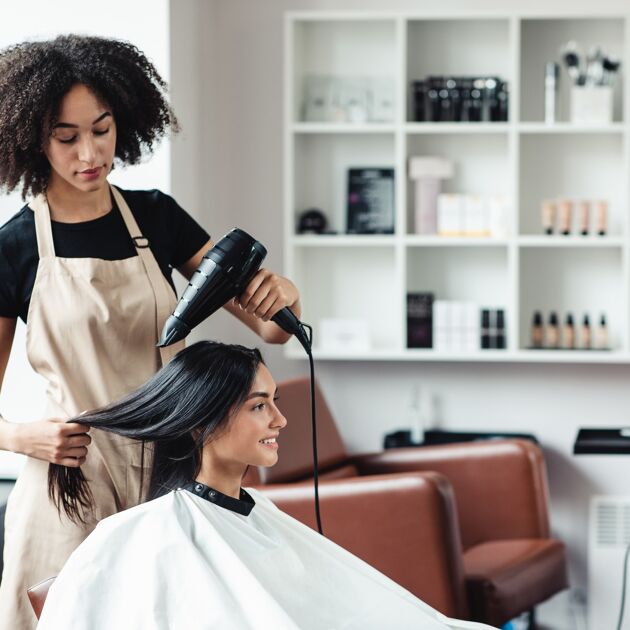 Vrouw met schort föhnt het haar van een lachende vrouw met donker haar.
