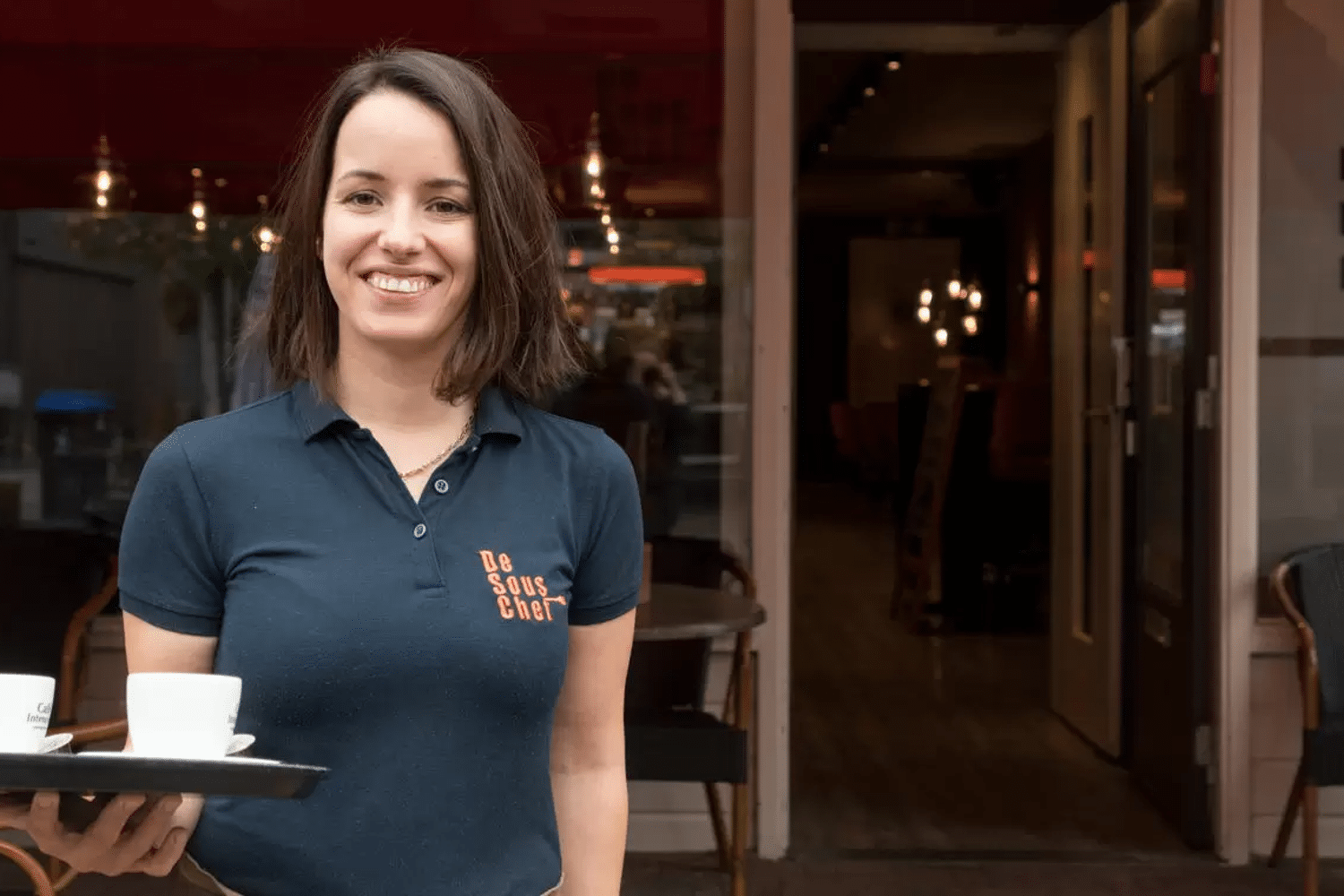 Vrouw met dienblad in handen staat lachend voor restaurant.