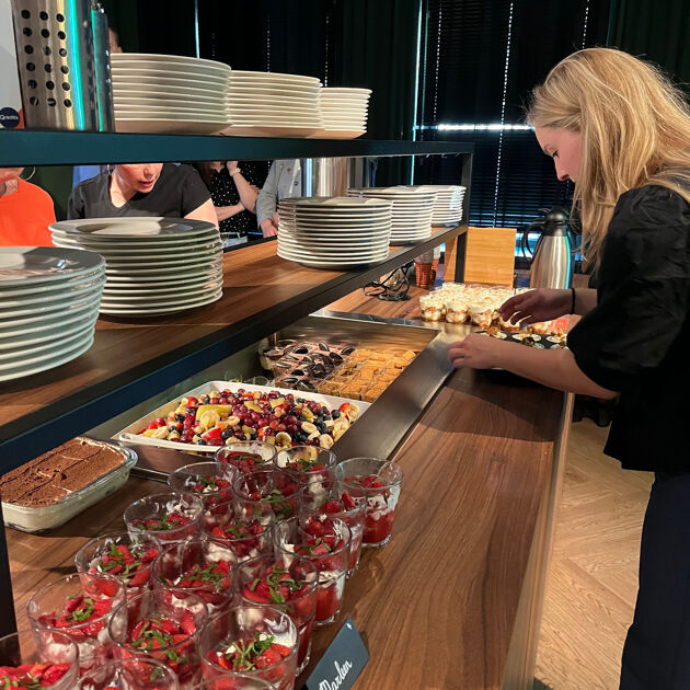 Een vrouw in zwarte kleding staat bij een tafel met allemaal eten en borden.