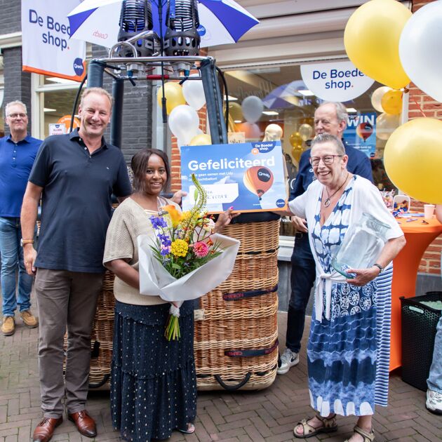 Twee vrouwen staan voor een luchtballon. Iemand heeft een bos bloemen vast en iemand anders een grote kaart.