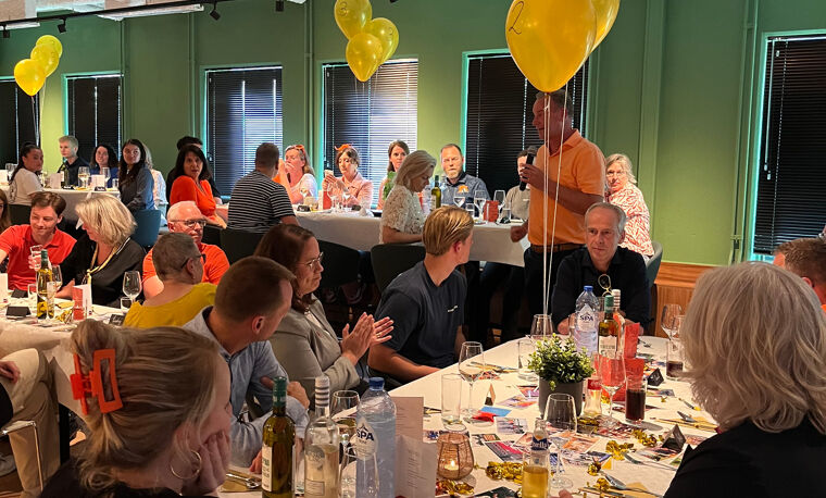 Een zaal vol mensen die aan tafels zitten met gele ballonnen die er boven hangen. 