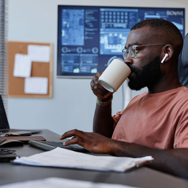 Man met een bril drinkt uit een kopje achter zijn bureau met computers.