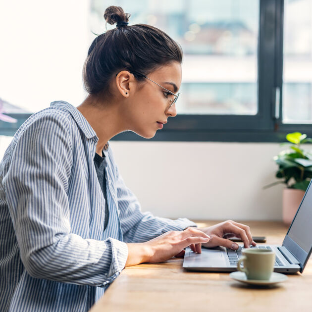 Vrouw met bril op zit aan tafel en kijkt naar laptop.