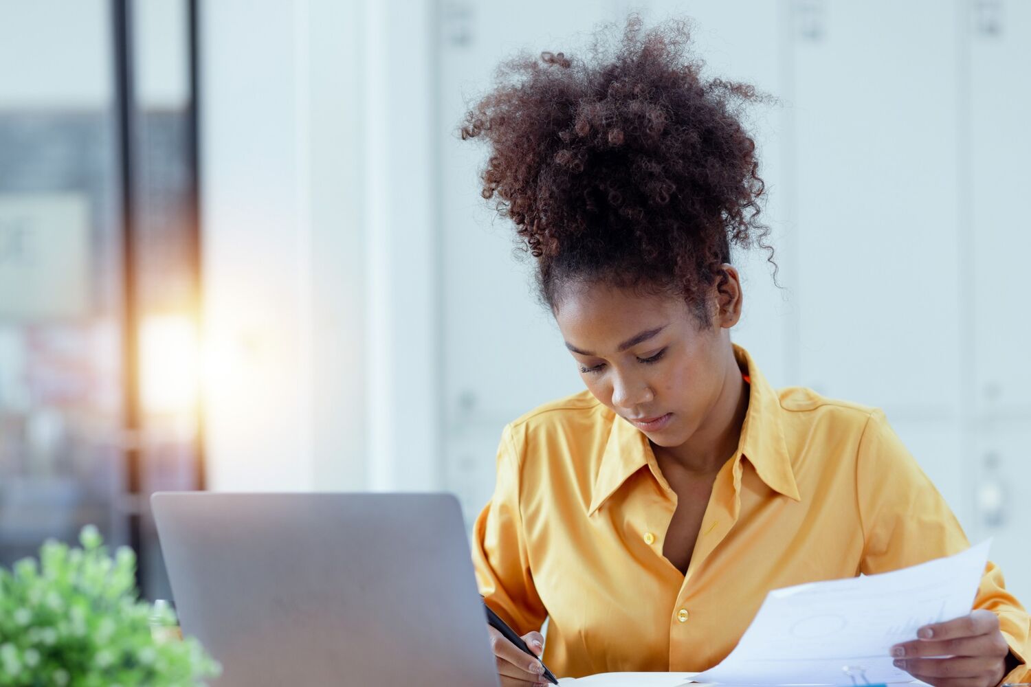 Een vrouw met een gele blouse schrijft op papier achter een laptop.