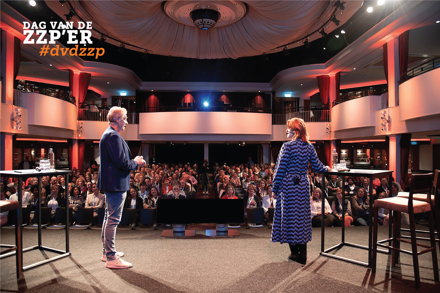 man en vrouw spreken op podium voor volle zaal