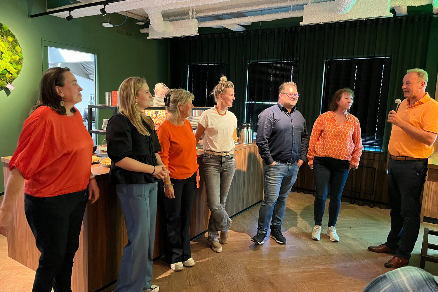 Een groep mensen staan op rij en kijken naar een man met een microfoon. Ze staan binnen in een ruimte die groen en bruin is.